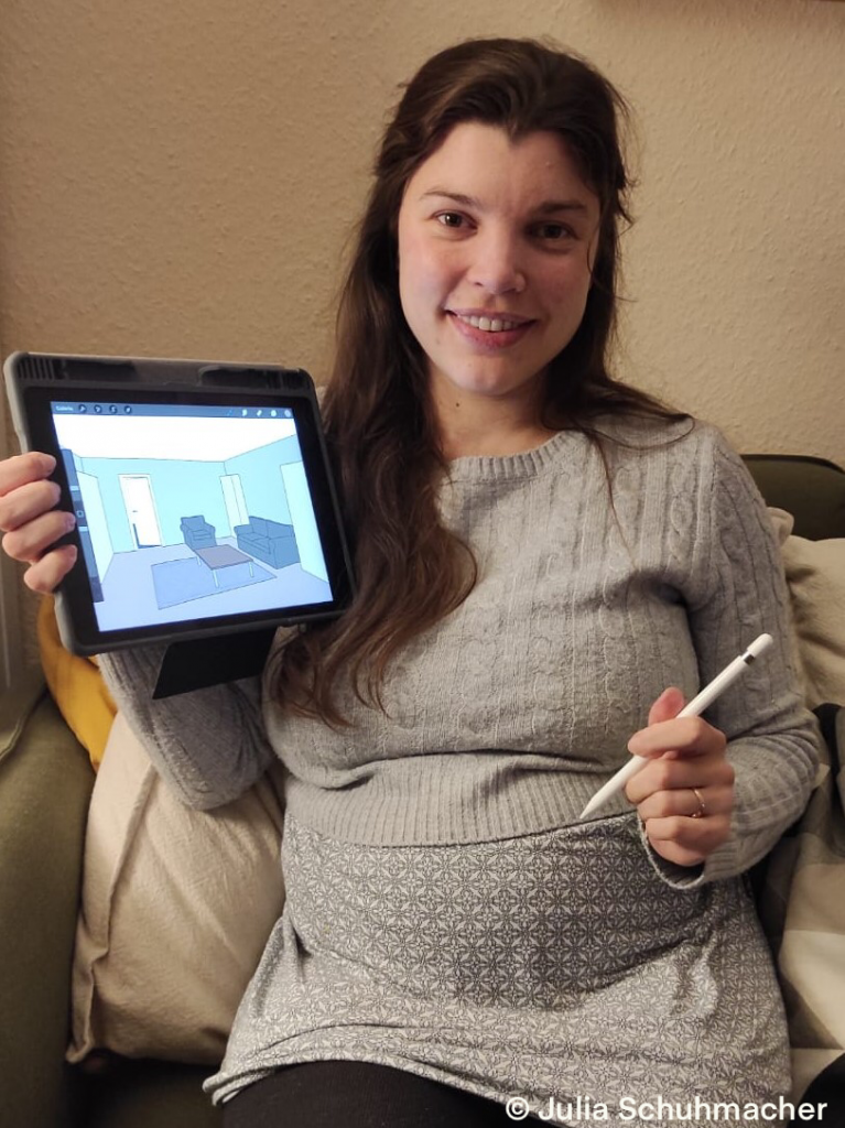 Auf dem Bild ist Julia Schuhmacher mit einem iPad und Pen zu sehen. Auf dem Tablet ist der Entwurf eines Raums aus dem Spiels zu sehen.